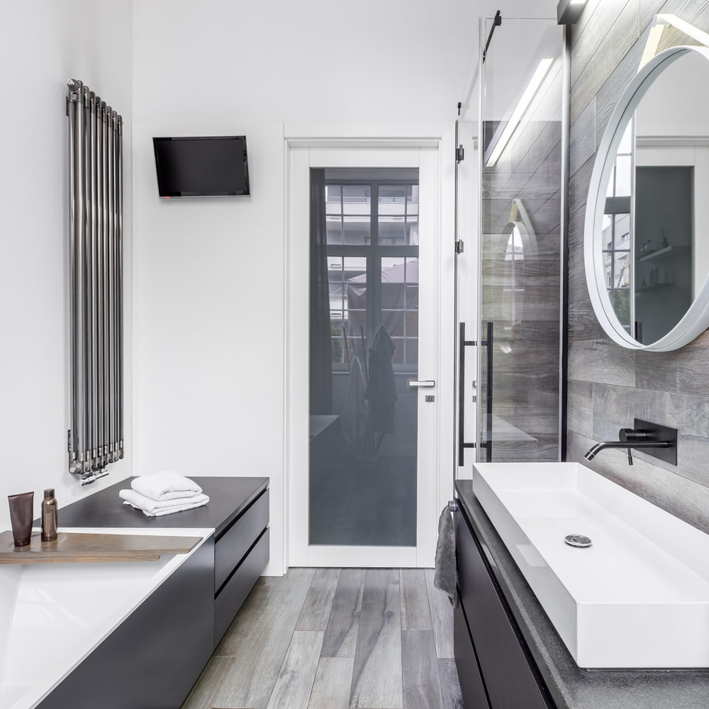 Bathroom with white glass door and sink at the right with round mirror on wooden flooring