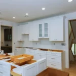 Carpenter fitting white crown molding above kitchen cabinets with a miter saw and finish nailer.