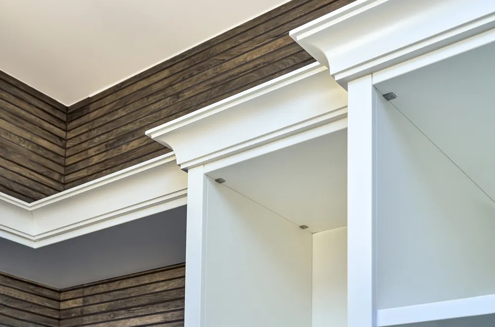 Carpenter fitting white crown molding above kitchen cabinets with a miter saw and finish nailer.