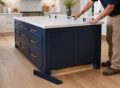 Contractor anchoring kitchen island base cabinets to wood cleats fastened into the structural subfloor.