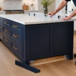 Contractor anchoring kitchen island base cabinets to wood cleats fastened into the structural subfloor.
