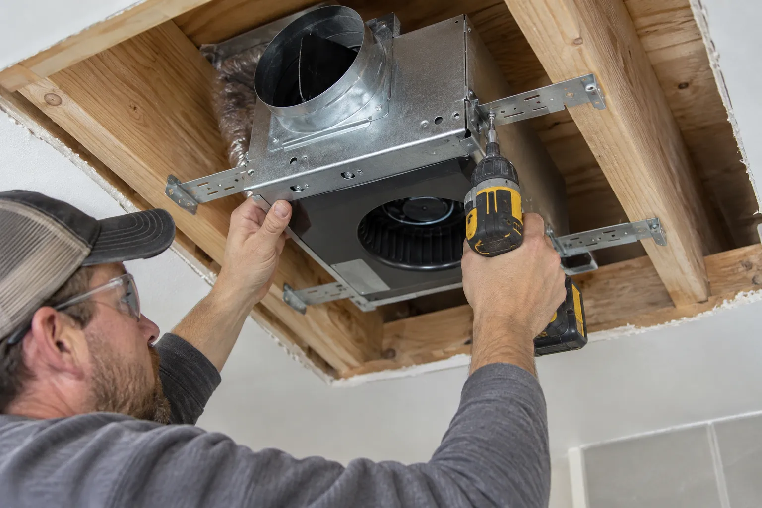 Contractor measuring and marking a ceiling opening for a bathroom exhaust fan near a glass shower.
